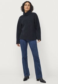 Geweven marineblauwe coltrui met een gestructureerd patroon, relaxte pasvorm, lange mouwen en zijsplitten, gecombineerd met donkere blauwe flared jeans en enkellaarsjes.