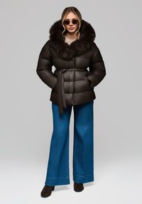 Giubbotto soffice marrone con cappuccio foderato in pelliccia, vita con cintura e motivo trapuntato, abbinato a jeans blu a gamba larga e stivali marroni scuri.