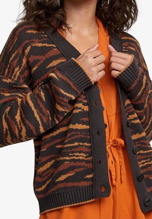 Kvinde iført en brun og orange stribet cardigan over en orange knappet kjole med bånd i taljen, der holder cardiganen nær brystet.