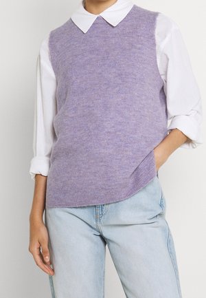 Person iført en lavendelstrikket ærmeløs sweater over en hvid skjorte med optrukne ærmer og lyseblå jeans, hånd i lommen.