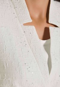 Witte textuurjacket met een V-hals en korte kraag, versierd met kleine zilveren pailletjes aan de voorkant. Gelaagd over een witte top.