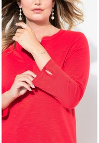 Rotes Langarm-Oberteil mit rundem Ausschnitt und Knopfdetail am Bündchen. Glatte Textur, lockere Passform und leicht lässige Silhouette.