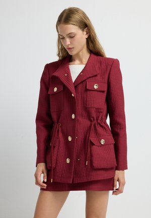 Veste matelassée rouge avec une taille ceinturée, boutons dorés, poches poitrine doubles et col à revers. Le tissu texturé ajoute de la profondeur.
