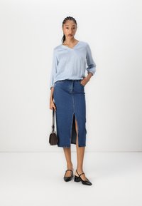 Camicia blu chiaro a maniche lunghe, con scollatura a V e piccoli pois bianchi, abbinata a una gonna in denim a vita alta con uno spacco frontale e scarpe nere con tacco.