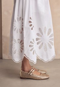 Baltas, išsiuvinėtas sijonas su gėlių išpjovomis ir banguotu apvadu. Metalinės aukso spalvos balerinos su pintu raštu ir dviem čiurnos dirželiais.