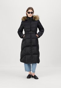 Juodas pūkinis paltas su dirbtinio kailio apdaila ant kapšos, dirželiu liemenyje ir užtrauktuko uždarymu. Paltas turi deimanto formos siuvinėjimą ir šonines kišenes.