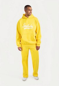 Żółta bluza z kapturem z przednią kieszenią, z białym logo w formie napisu. Do kompletu żółte spodnie dresowe i białe trampki.