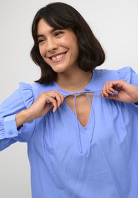 Blusa azzurro chiaro con scollatura a V e dettaglio con nodo all'collo. Caratterizzata da maniche a sbuffo e tessuto morbido e testurizzato. Polsini con bottoni.