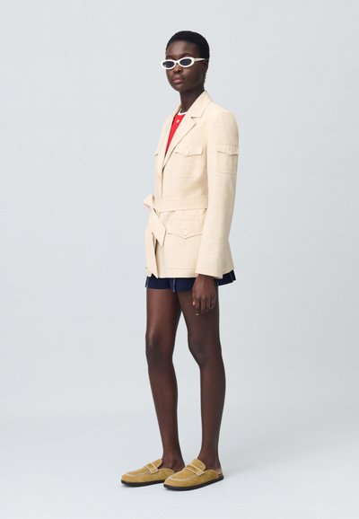 Blazer beige adjustable, associé à un haut rouge, un short en denim et des chaussures jaunes sans lacets. Le mannequin porte des lunettes de soleil blanches en forme d'oval.