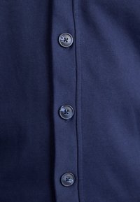 Närbild av marinblått tyg med tre blanka, fyrhåliga, runda knappar vertikalt placerade på en placket.