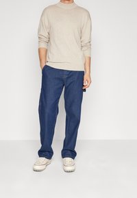 Mann, der einen beigen Rollkragenpullover, lockere dunkelblaue Jeans und weiße Turnschuhe trägt, mit einer Hand in der Tasche steht.