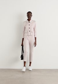 Lichtroze op maat gemaakte blazer met bruine knopen, bijpassende slim fit broek en witte sneakers; model houdt een kleine zwarte tas vast, staand op een neutrale achtergrond.