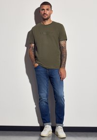 Muž s tetováním na celém rukávu, oblečený v olivově zeleném tričku, modrých nohavicích nažehlených do kotníků a bílých teniskách, stojí proti bílé zdi.