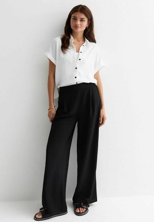 Witte kortelijst knoopshirt met zwarte knopen, gecombineerd met zwarte wijde broek en zwarte sandalen met zilveren accenten.