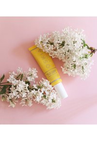 Gul tub av Oskia SPF 30 Vitamin Face Cream på en rosa bakgrund, omgiven av kluster av vita blommor, slät yta, ogenomskinlig kork.