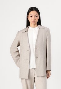 Beige takasella bleiseri, jossa on taitettu kaulus, yksittäinen napitus ja kaksi taskua, yhdistetty valkoisen ribbivillapaidan kanssa. Sujuva, tekstuuriinen kangas.