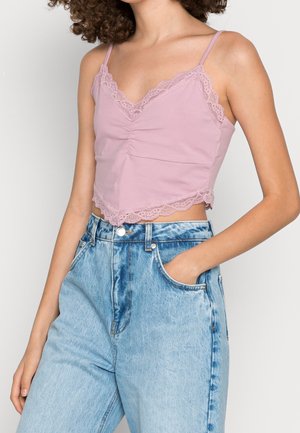 Frau trägt ein fliederfarbenes, mit Spitze besetztes, kurzes Camisole und hoch taillierte hellblaue Jeans, die eine Hand in der Tasche hat.