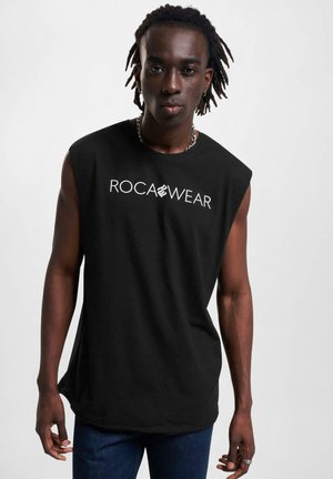 Svart ermeløs t-skjorte med hvit "ROCA WEAR" logo, mykt bomullsmateriale, avslappet passform og rund nederkant, kombinert med mørkeblå jeans.