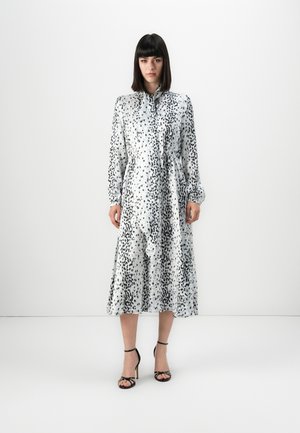 Woman wearing a long-sleeve white midi dress with black irregular spots, standing against a plain white background, wearing black high-heel sandals.