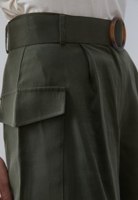 Zbliżenie na osobę w oliwkowych spodni z wysokim stanem, z boczną kieszenią na klapę i szerokim paskiem z dużą okrągłą klamrą.