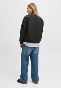 Zwarte bomberjack met geribde manchetten en zoom, gedragen over een lichtblauw gestreept shirt, gecombineerd met losse blauwe jeans, vanuit de achterkant bekeken.