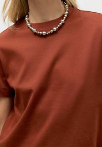 Rustfarvet bomulds t-shirt med rund hals, der har en glat tekstur. Halsen er prydet med en sølvfarvet perlehalssmykke for ekstra detaljer.