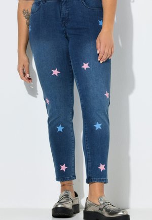 Persoon die blauwe jeans draagt, versierd met roze en blauwe sterren, zilveren plateau loafers en een kleine tatoeage op de linker onderarm.