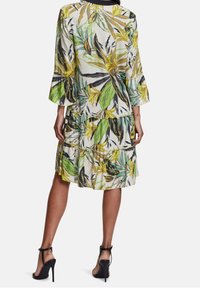 Robe à imprimé floral avec un design à étages, des manches longues en forme de cloche et une longueur de genou. Les couleurs incluent le vert, le jaune et le noir.