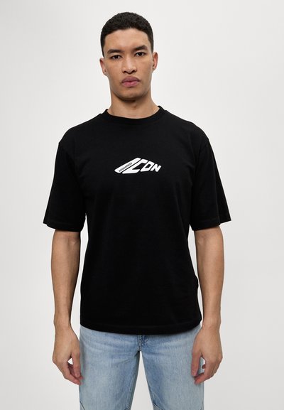 T-shirt en coton noir avec un col ras du cou et des manches courtes. Présente un logo blanc prominent au centre. Porté avec un jean bleu clair.