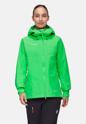 Frau trägt eine hellgrüne wasserdichte Jacke mit Kapuze und schwarze Hose, steht und schaut nach vorne vor weißem Hintergrund.