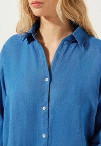 Blå linneskjorta med knäppning fram, krage och lätt skrynklig textur. Har små vita knappar längs placket.
