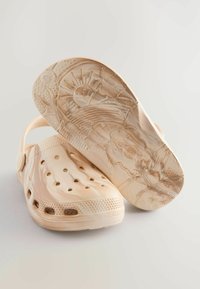 Ljusbeige träskor med ventilationshål och en bakrem. Sulan har präglade motiv av sol och moln, strukturerad för grepp.