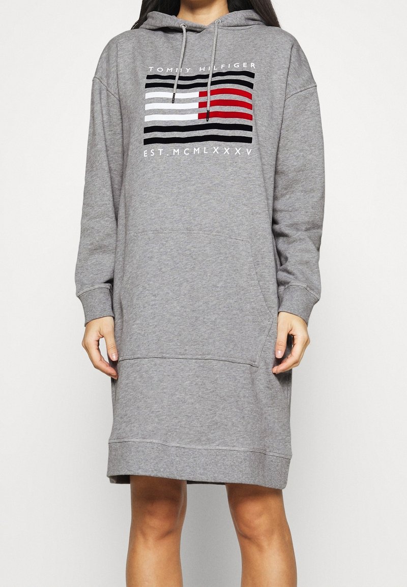 Femme portant une robe à capuche grise Tommy Hilfiger avec une poche frontale et un logo avec des rayures noires, blanches et rouges sur la poitrine.