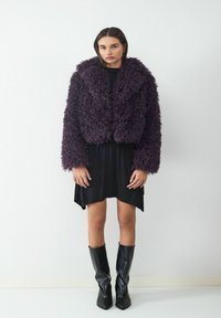 Fioletowa kurtka z futra syntetycznego, krótka, z kołnierzem typu szal, noszona na czarnej sukience, w połączeniu z czarnymi kozakiem do kolan. Teksturowany i oversize'owy fason.