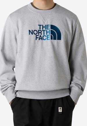 Osoba nosząca jasnoszary sweter z niebieskim logo "The North Face" i czarne spodnie, z rękami w kieszeniach.