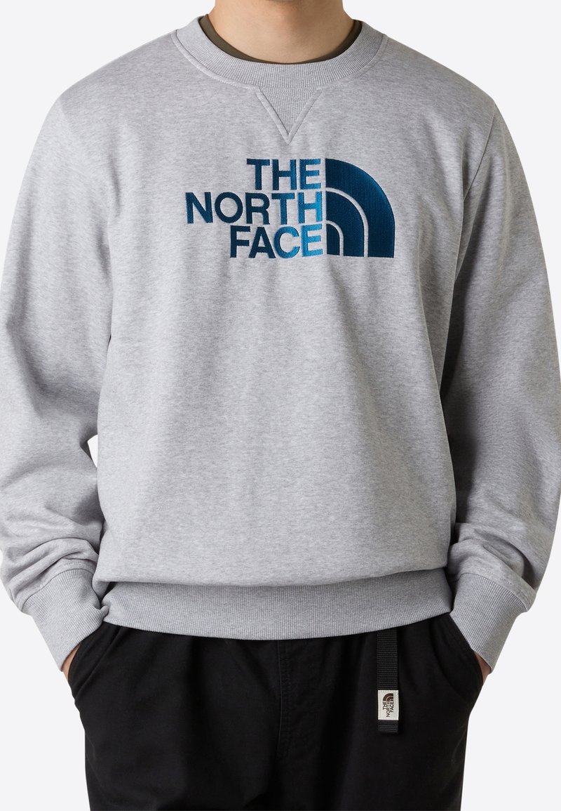 Personne portant un sweat-shirt gris clair avec un logo bleu « The North Face » et un pantalon noir, les mains dans les poches.