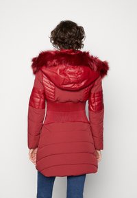 Chaqueta de plumón roja con capucha forrada de pelo, cintura acanalada y diseño de hombros acolchados. Textura suave con secciones acolchadas en contraste.