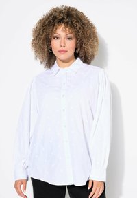 Camisa blanca de botones con patrones de corazones sutiles, mangas largas con puños fruncidos y un cuello clásico.