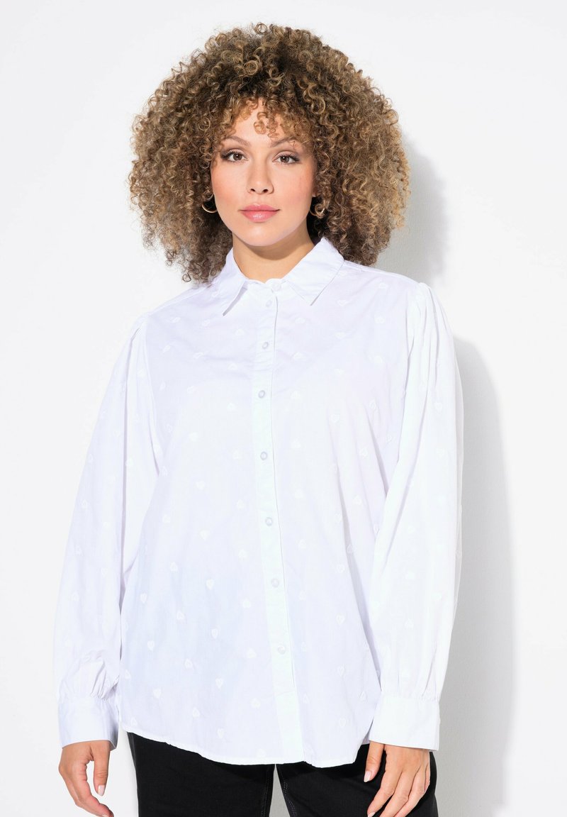 Camisa blanca de botones con patrones de corazones sutiles, mangas largas con puños fruncidos y un cuello clásico.