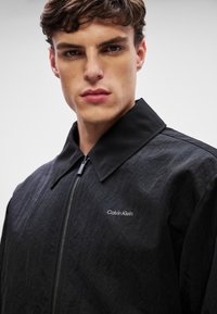 Sort zip-up jakke med en stor krave, lavet af tekstureret stof. Har et hvidt "Calvin Klein" logo på venstre side.