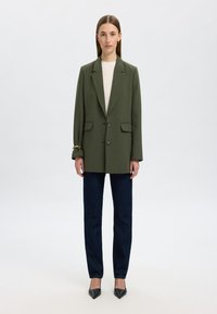 Olivenfarget oversized blazer med klaffelapper, to frontlommer og knappelukking, kombinert med mørkeblå rette bukser og svarte høyhælte sko.