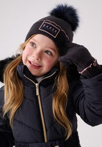 Nuori tyttö pukeutuneena tummansiniseen takkiin, neulottuun hattuunsa, jossa on tupsu, ja sopiviin hansikkaisiin, hymyillen ja säätäen käsillään hattuaan yksiväristä taustaa vasten.