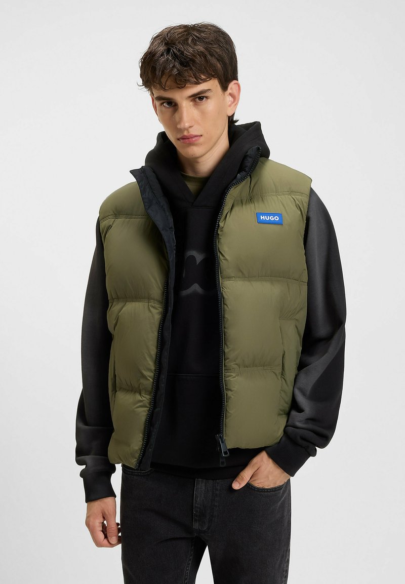 Chaleco acolchado verde con cuello alto, cremallera frontal y bolsillos laterales; cuenta con un parche de logo "HUGO" azul en la parte superior izquierda.