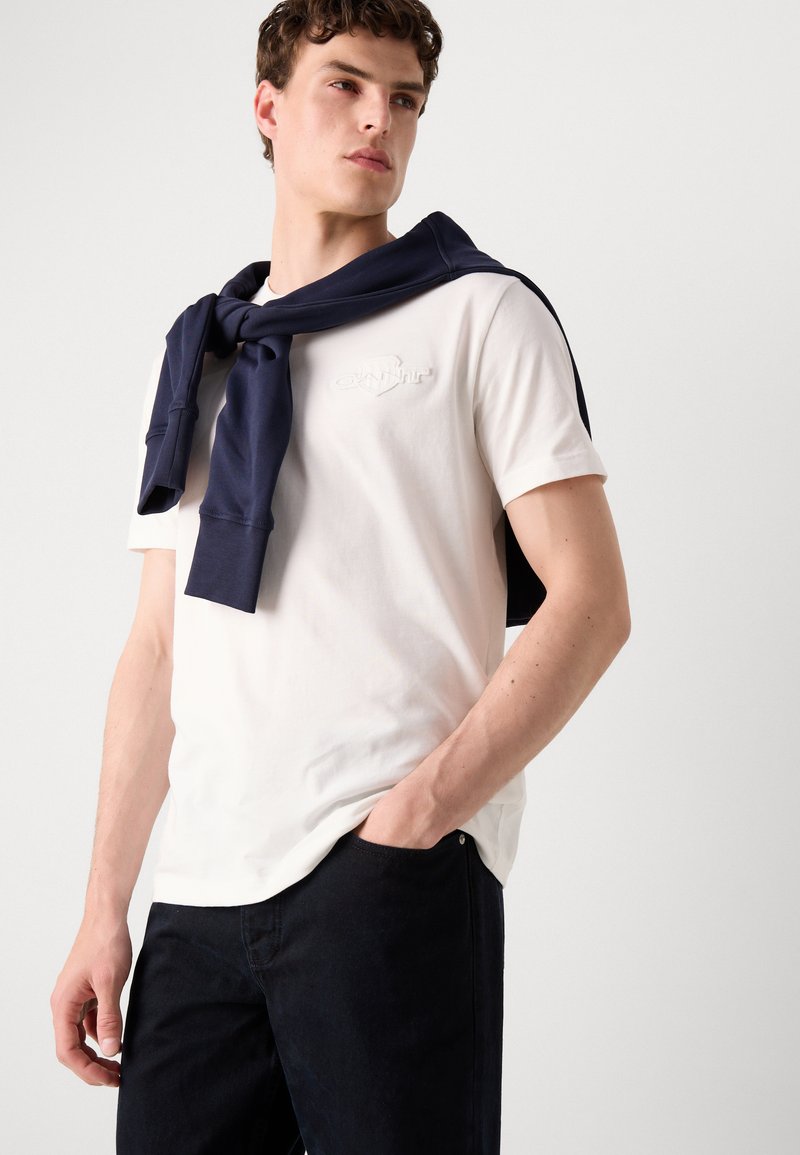 Weißes Baumwoll-T-Shirt mit gesticktem Logo, kombiniert mit einem dunkelblauen Pullover, der am Hals gebunden ist, und dunklen Jeans. Einfarbiger Hintergrund.