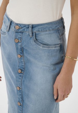 Lyseblå denimnederdel med knapper foran og lommer, båret med hvid top, accessoriseret med guldbøjle og ring på venstre hånd.