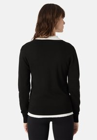 Černý pletený svetr s kulatým výstřihem a ribovanými manžetami, pod nímž vykukuje bílá košile s odřeným lemem. Jednoduchý design bez vzorů.