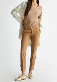 Pull en beige clair avec un col en V, cardigan tricoté crème, pantalon marron clair fuselé avec des boutons sur le côté, et mocassins dorés. Accents en tissu texturé.