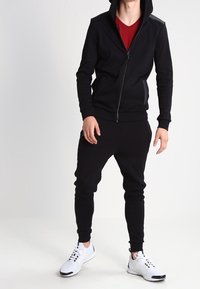 Czarna bluza na suwak z wypukłym akcentem na ramieniu, w zestawie z dopasowanymi czarnymi dresami i białymi butami sportowymi. Widoczna czerwona podkoszulka.