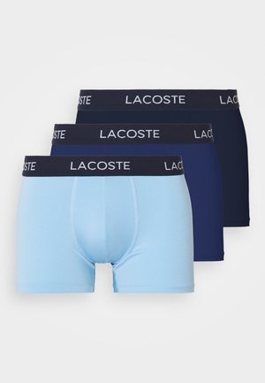 Korte bomullsboxere i lys blå og to nyanser av marineblå, med et mørkt liv med "LACOSTE"-logo i hvitt.