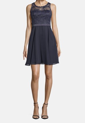 Vestido sin mangas azul marino con cuerpo de encaje y falda fluida, combinado con tacones negros con tiras en el tobillo, en una modelo femenina de pie.